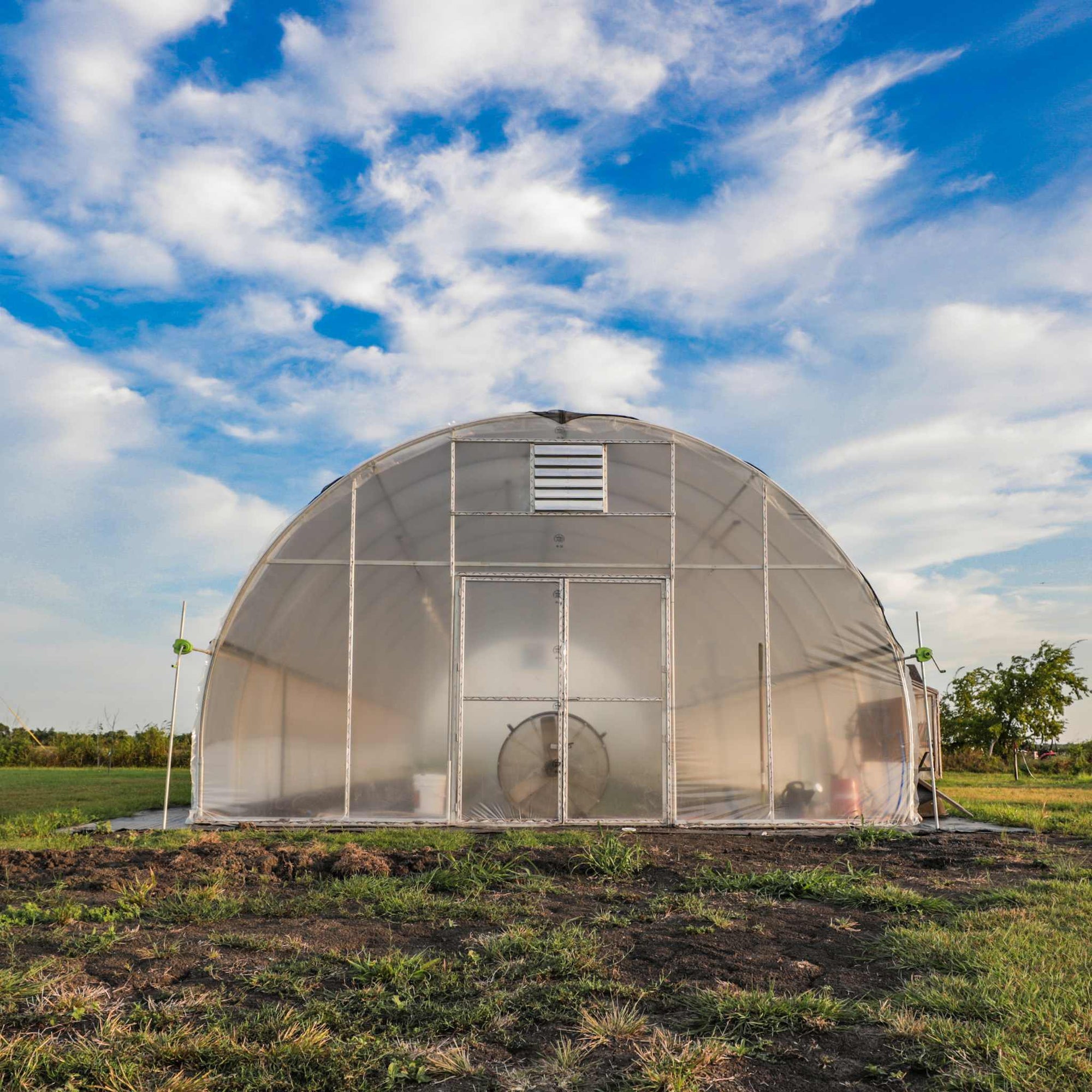 A sturdy USA-manufactured round hoop house with a vented front door, clear greenhouse plastic covering, and durable metal framing, set against a vibrant blue sky with scattered clouds. Ideal for professional and homestead growers seeking longevity and efficiency.